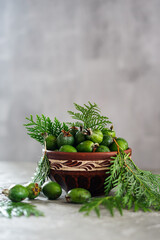feijoa fruits in a brown clay plate with scattered feijoa fruits on a gray background with thuja leaves vertical photo
