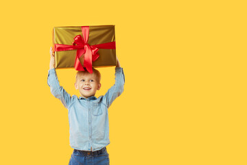 Portrait of happy cute child boy holding a large gold gift box above his head and looking at camera isolated on yellow background. Wow face