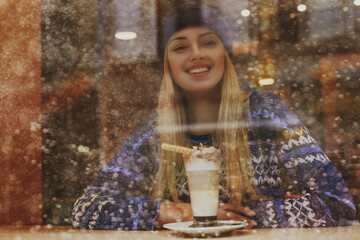 Fototapeta na wymiar Christmas, winter holidays conception: happy smiling woman drinks hot drink in cafe. View through the window glass. Snowfall. Copy, empty space for text