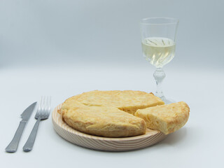 Tortilla de patata sobre un plato de madera, cuchillo y tenedor y un vaso con vino en fondo blanco.