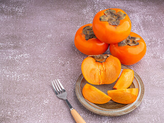 Fresh ripe persimmons with slices of persimmon on a plate with cutlery and whole fruit on gray stone background. Space for text. Close-up photo. Concept of health fruit