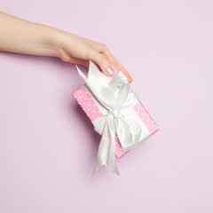 Pink gift with white silky ribbon in female hand on pastel lilac background. Minimal background composition card