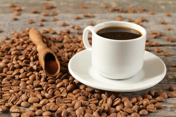The white cup of coffee is near the wooden scoop with ground coffee among the scattering of coffee beans on a table.