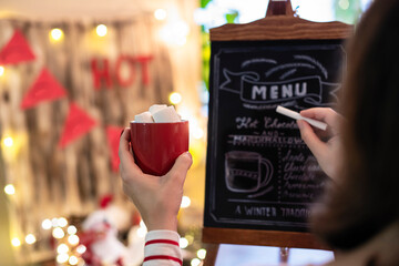 Woman with cup of cocoa with marshmallows drawing christmas menu of cocoa bar on small chalk board.