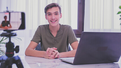 Man blogger videotapes his vlog at home. Teenage boy recording his video blog. Young man speaks in front of a video camera for his blog channel. Vlogger makes online streaming using smartphone