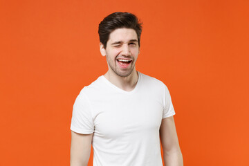 Blinking cheerful funny handsome attractive young man 20s wearing basic casual white blank empty t-shirt standing looking camera isolated on bright orange colour wall background, studio portrait.