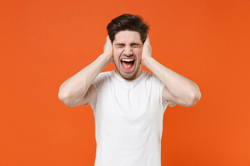Fototapeta na wymiar Crazy frustrated young man 20s in basic casual white t-shirt standing keeping eyes closed put hands on head screaming looking camera isolated on bright orange colour wall background, studio portrait.