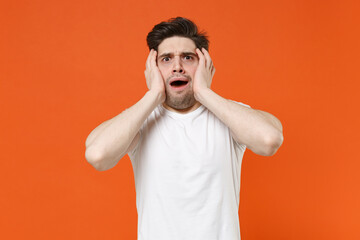 Fototapeta na wymiar Shocked worried scared young man wearing basic casual blank white t-shirt standing put hands on cheeks keeping mouth open looking camera isolated on bright orange colour background, studio portrait.