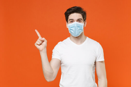 Young man wearing casual white t-shirt sterile face mask to safe from coronavirus virus covid-19 during pandemic quarantine pointing index finger up isolated on orange background studio portrait.