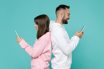 Side view of smiling young couple two friends man woman 20s in white pink hoodie standing back to back using mobile cell phone typing sms message isolated on blue turquoise background studio portrait.