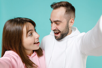 Close up of shocked young couple two friends man woman 20s in white pink casual hoodie standing doing selfie shot on mobile phone isolated on blue turquoise colour wall background studio portrait.