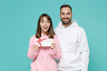 Funny laughing excited young couple two friends man woman 20s wearing white pink casual hoodie hold gift certificate looking camera isolated on blue turquoise colour wall background studio portrait.