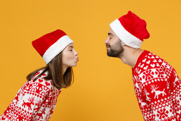 Side view of pretty young Santa couple friends man woman in sweater Christmas hat keeping eyes closed kissing isolated on yellow background studio. Happy New Year celebration merry holiday concept.