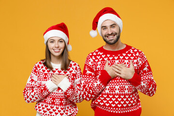 Smiling young Santa couple friends man woman in red sweater Christmas hat holding hands folded on heart isolated on yellow background studio portrait. Happy New Year celebration merry holiday concept.