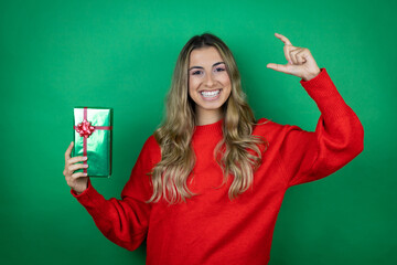 Young beautiful girl holding gift over isolated green background smiling and confident gesturing with hand doing small size sign with fingers . Measure concept.