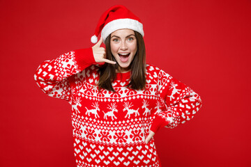 Excited young brunette Santa woman 20s in sweater Christmas hat doing phone gesture like says call me back isolated on red background studio portrait. Happy New Year celebration merry holiday concept.