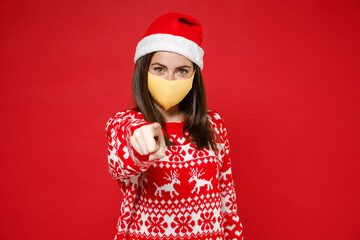 Young Santa woman in sweater Christmas hat face mask to safe from coronavirus virus covid-19 point index finger on camera isolated on red background. Happy New Year celebration merry holiday concept.