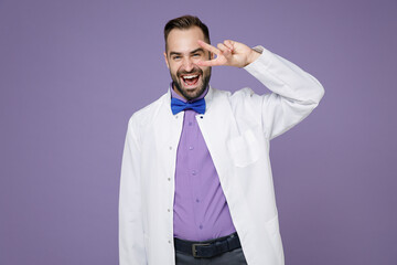 Cheerful young bearded doctor man wearing white medical gown showing victory sign looking camera isolated on violet colour background studio portrait. Healthcare personnel health medicine concept.