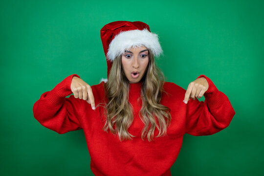 Young beautiful girl wearing Christmas Santa hat holding gift over isolated green background surprised, looking down and pointing down with fingers and raised arms