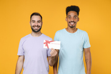 Smiling young two friends european african american men 20s wearing violet blue casual t-shirts hold gift certificate looking camera isolated on bright yellow colour wall background studio portrait.