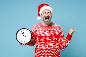 Fototapeta na wymiar Happy young Northern bearded man frozen face in Santa hat Christmas sweater hold clock doing winner gesture isolated on blue background. Happy New Year celebration merry holiday winter time concept.