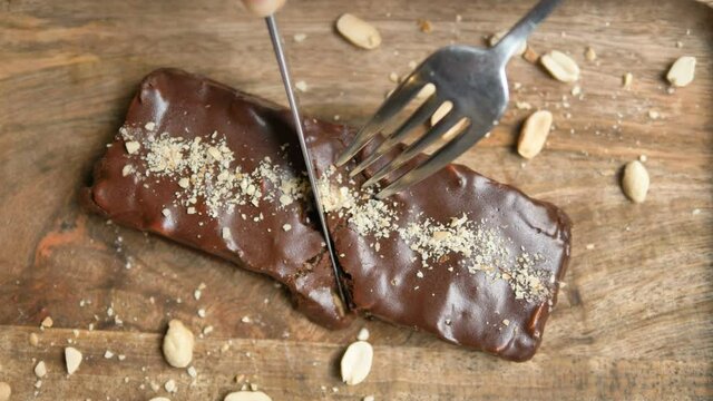 Fork and knife cutting delicious dessert gluten free caramel bar with peanuts lying on wooden board extreme closeup slow motion
