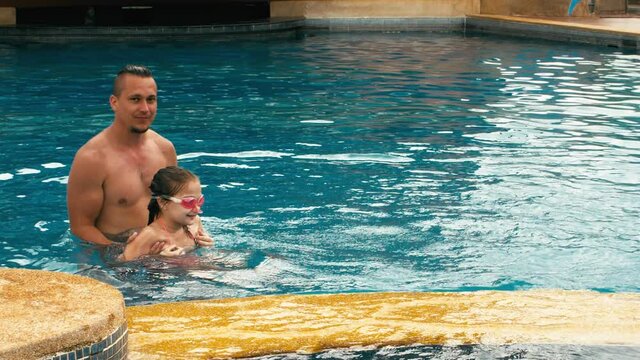 The father with little daughter have fun in the pool. Dad plays with the child. The family enjoy summer vacation in a swimming pool jumping, spinning, splash water. Slow motion.