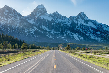 Straight road towards Grand Teton mountains at Grand Teton National Park, United States. Green tress and some light rays can be see coming from the left