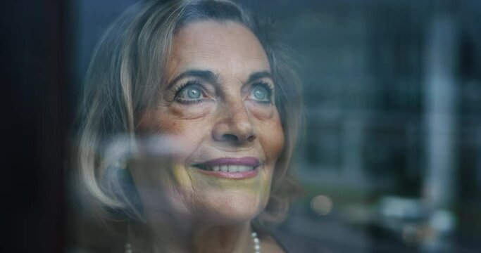 Cinematic close up of an elderly smiling woman is looking out window of her house in the morning.