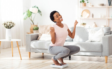 Excited black woman sitting on scales at home, happy with result of her slimming diet, copy space