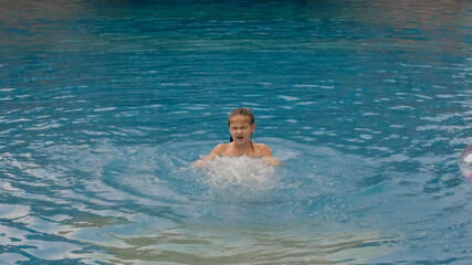 The little cute girl have fun in the pool. The child enjoy summer vacation in a swimming pool jumping, spinning, splash water. Happy childhood.