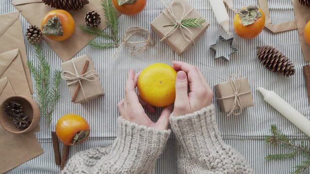 A girl in a knitted sweater holds a tangerine in her hands, surrounded by Christmas gifts. Christmas concept, new year's holiday atmosphere. Craft paper gift wrapping handmade.