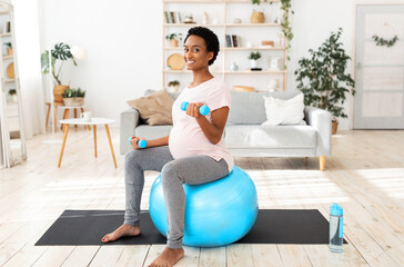 Happy black pregnant woman working out with dumbbells on fitball at home