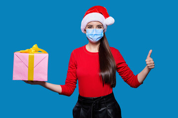 Fototapeta na wymiar Woman wearing protective medical mask and Santa hat holding pink gift box, showing thumb up gesture isolated on blue background. New year gift during coronavirus.