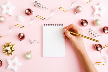 Christmas decorations, Woman's hand writing in blank Notebook on pastel pink background. New Year composition. Flat lay, top view, copy space.