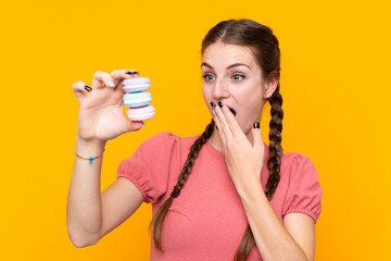 Young woman over isolated yellow background holding colorful French macarons and with surprise expression