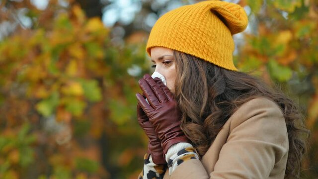 Hello november. modern woman in beige coat and orange hat with napkin blowing nose outdoors in the city park in autumn. 
