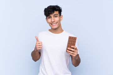 Young Argentinian man over isolated blue background taking a chocolate tablet and with thumb up