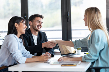 Attractive real-estate agent shaking hands with young couple after signing agreement contract in...