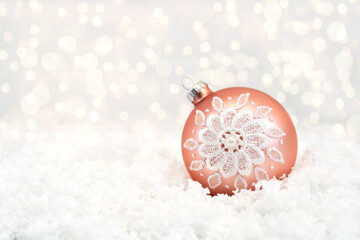 A rose gold Christmas glass ball with a white pattern lies on white snow. Bokeh in the background. There is some free space on the left for text. Close-up