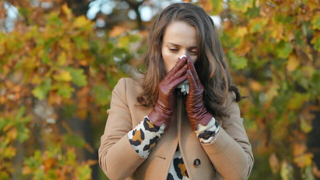 Hello autumn. modern 40 years old woman in brown coat and yellow hat with napkin blowing nose outdoors in the city park in autumn. 
