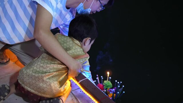 4K Asian child boy and father floating banana leaf krathong with candle lighting at pond in Loy Krathong festival day. Happy family activity together outdoor.