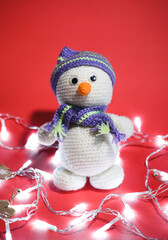 A knitted white snowman with a carrot for a nose sits on a red Christmas background with lights and decorations. Charming snowman toy with knitted hat and scarf, handmade.