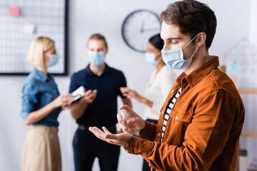 Fototapeta na wymiar manager in medical mask spraying disinfectant on hands near colleagues on blurred background