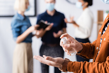 Obraz premium cropped view of businessman spraying sanitizer on hands while colleagues talking on blurred background