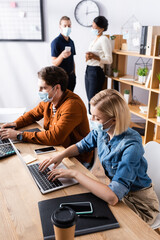 Fototapeta premium managers in medical masks working on laptops near multicultural colleagues talking on blurred background