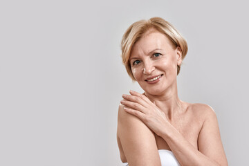 Portrait of beautiful middle aged woman smiling at camera, touching her shoulder, posing isolated against grey background. Beauty, skincare concept