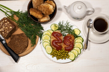 There is lunch for one person on the table. On a white plate are cutlets with tomato sauce, green peas, pieces of cucumber and a sprig of dill. There is a tea set and a board with bread nearby.
