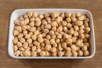 A white ceramic dish full of raw white chickpeas with a yellowish tone after being soaked on a wooden table.
. Organic textures as a background. Vegan food and legumes.