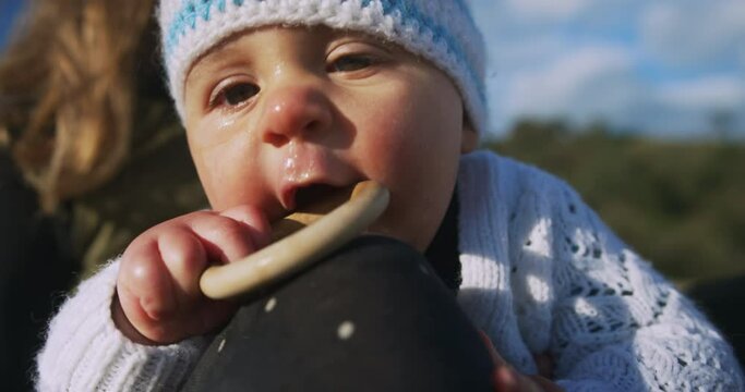 A little baby chewing on a wooden ring is sitting on the beach with his mother in the autumn sun
